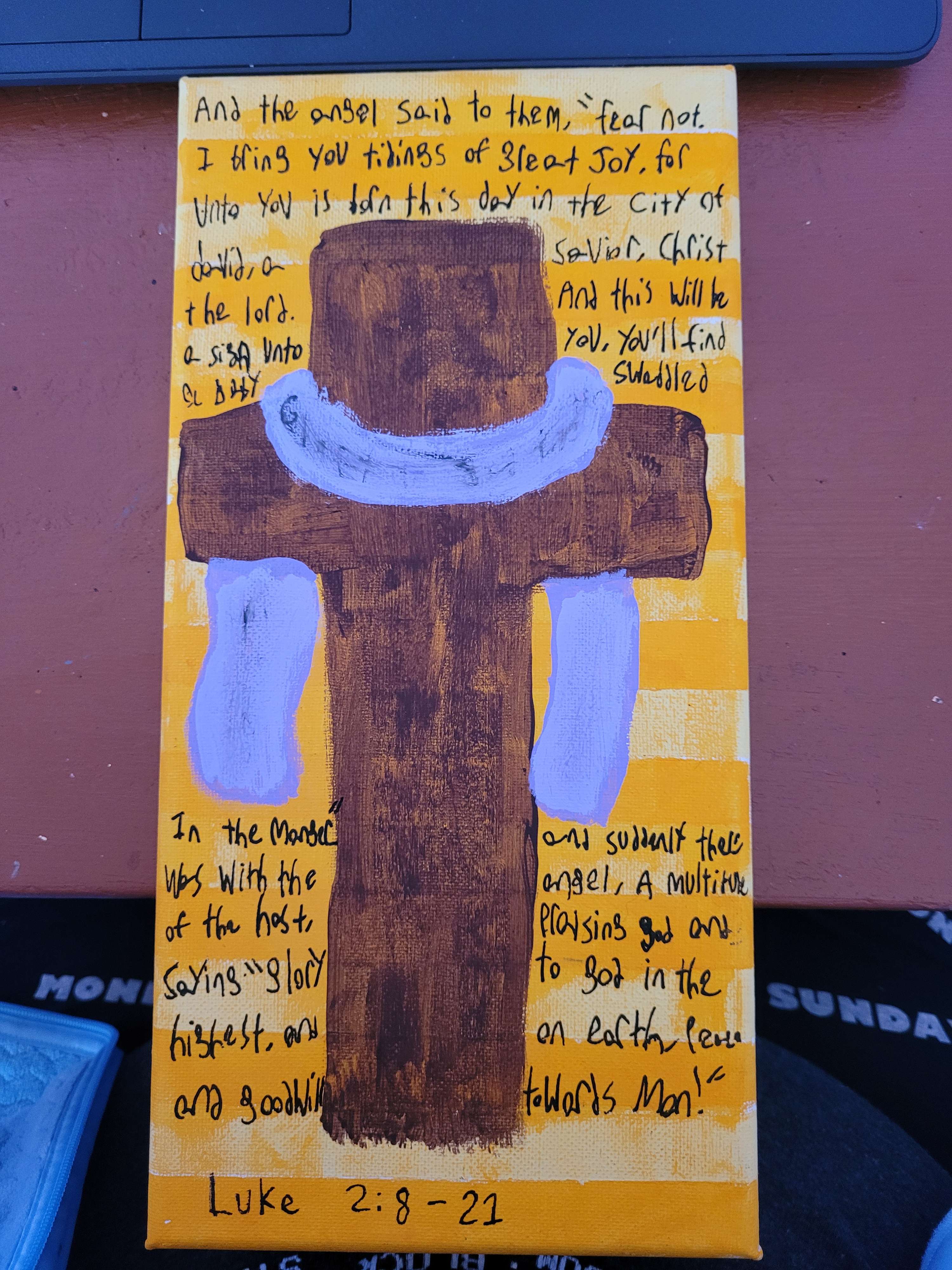 Painting of a cross with purple fabric on it, a bible quote of the christmas story is written on it.